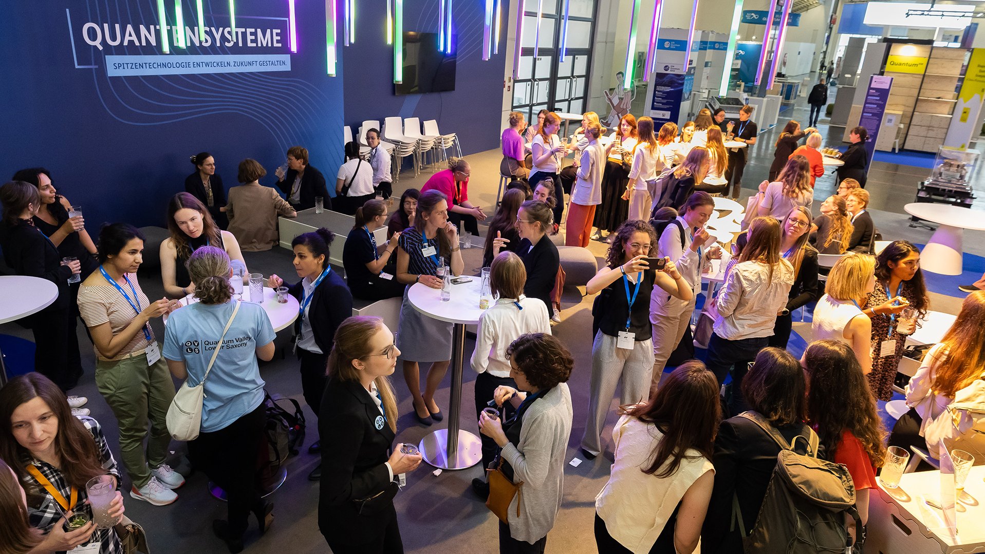 Eine große Gruppe Frauen feiert an einem Messestand eine Feier, zu sehen sind Stehtische und der Schriftzug Quantensysteme
