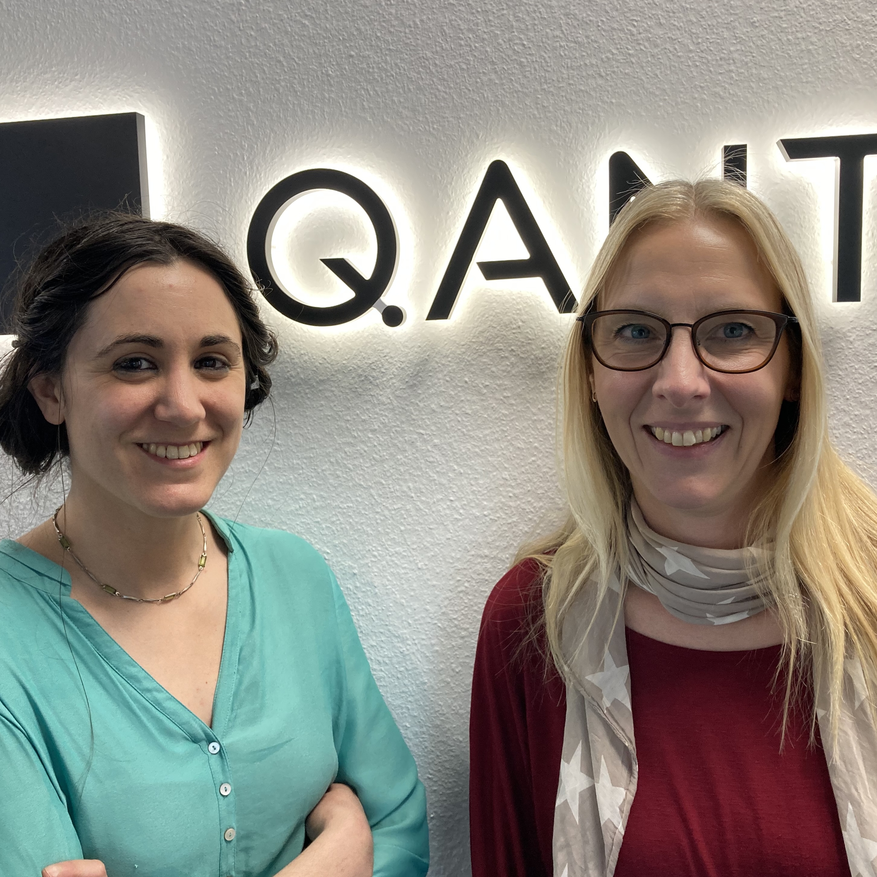 Portrait von Lara Torralbo-Campo und Sandra Mebben vor dem Logo der Firma Q.ANT